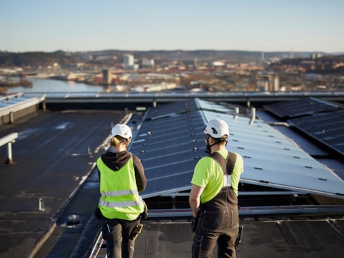 Two workers with bright yellow safety vests and helmets seen from behind looking at solar panels installed on the roof of a building